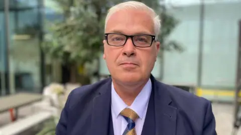 Michael Hudson is looking directly at the camera, unsmiling. He is wearing a blue three-piece suit, with yellow and blue tie. He has black glasses and white hair. He is stood in a glass atrium.