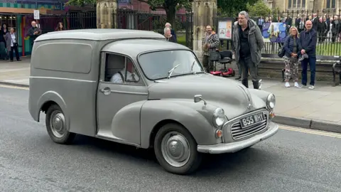 An old-fashioned grey 1964 Morris Minor van is being driven along a road.