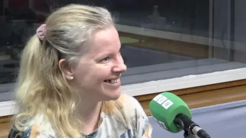A blonde woman is smiling next to a green microphone. She is sitting in a studio and is wearing a white, blue and yellow top.