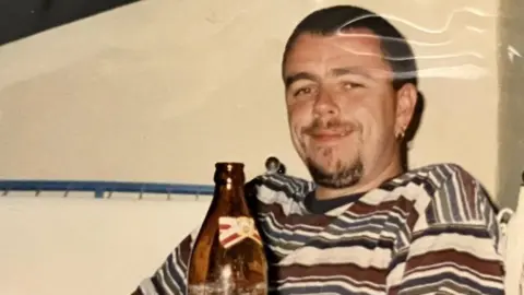 Alison Mark Jenner, pictured by Alison, on one of their holidays. He wears a striped t-shirt and a beer bottle sits on the table in front of him.