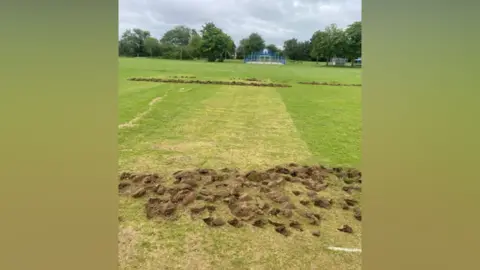 Westcroft Cricket Club A long line of freshly uprooted turf stretching across the middle of a cricket pitch. Large clumps of soil are scattered in a continuous strip, sharply contrasting with the surrounding green grass. In the background, trees, a fenced sports area, and distant houses line the edge of the open space. The cloudy sky casts a dull, even light over the scene, emphasizing the rough, disturbed ground.
