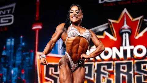 gilcoproductions A female bodybuilder wearing a union jack bikini poses on a stage, with a colourful backdrop saying Toronto. She is smiling widely, and has long black hair down to her waist, and diamante bracelet and earrings.
