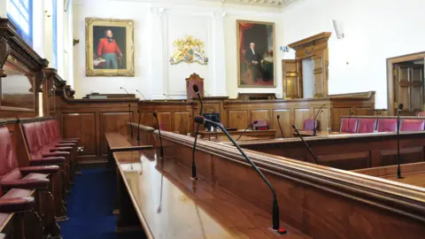 Guernsey States' debating chamber. A wood-panelled room with oil paintings, multiple red leather-upholstered seats and tables, plus several large windows.