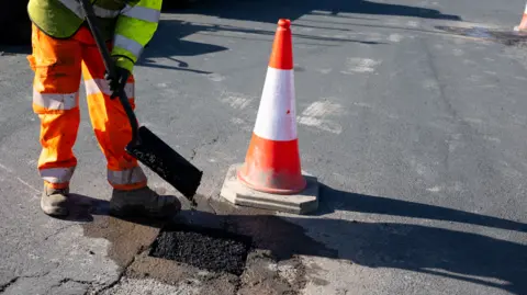 A man in orange reflective trousers and a yellow jacket holding a shovel on a road next to an orange traffic cone and a square of fresh tarmac