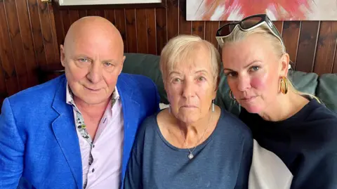 Aldo Zilli, a bald man wearing a blue jacket and light-coloured shirt, sitting next to his mother-in-law Maureen in the centre, and his wife Nichola on the right. They are all sitting on a green leather sofa