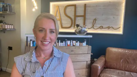 Ellie Pack is on the left. She is smiling and has her blonde hair pulled back. She is wearing a striped short-sleeved button-up shirt and a gold necklace. Behind her is a brown sofa and a sign which says Ash Salon on it written in gold.