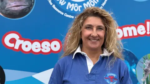 Standing in front of a blue sign that says welcome in English and Welsh. she has long curly blonder hear and is wearing a blue button down shirt with the attraction logo on the chest. 