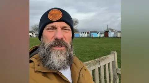 Supplied A screenshot of a selfie-style video of a man with a beard wearing a hat and a tan coloured jacket. He is sitting on a bench on a greensward. In the background there are rows of colourful beach huts.
