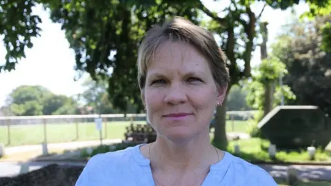 A woman with short blonde hair and a light blue blouse on smiles at the camera. Behind her is a green tree and grassed area, with sunlight streaming down. 