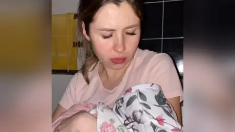 Maryna Korolova Maryna holding her newborn baby. The baby is in a floral onesie and the young woman is in a pink pyjama top. 