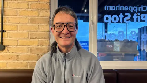 James smiling while sitting on a brown leather bench. Behind him is a honey coloured brick wall and large window that looks out onto the car park. James wears a grey zip up fleece and wears large black framed glasses.