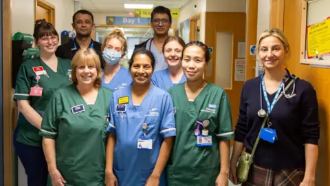 Salisbury District Hospital A group of nurses and doctors wearing green and blue scrubs are posing for a picture in the middle of a hospital ward. There are nine people in total wearing lanyards, and one is wearing a face mask.