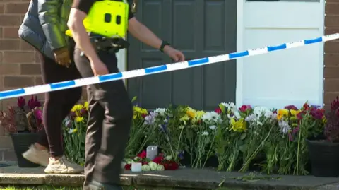 A member of the public and a police officer walk past floral tributes that have been left for Jahziah Coke and are lined up against a wall.