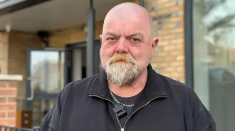 Paul is photographed standing outside his new home. He is bald but has a grey goatee beard. He is wearing a black zipped top.