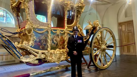 Provided The image shows a uniformed police officer stood in front of the gold state coach. The carriage is gold plated with large wheels and hand painted people on the doors.