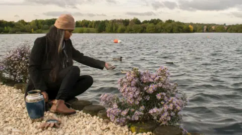 A woman in a dark jacket is seen pouring what appear to be ashes into a body of water. Alongside if an urn typically used to hold ashes