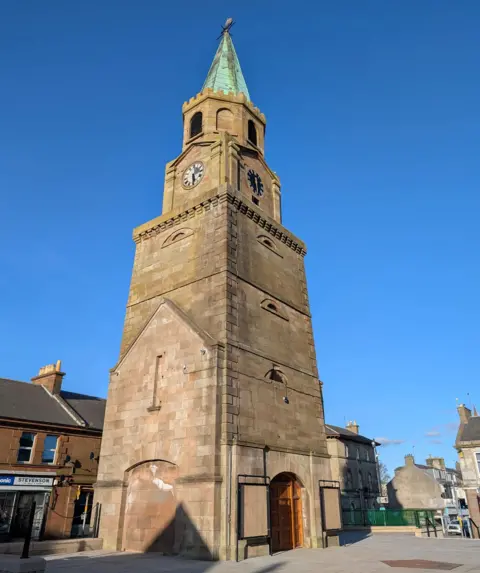 Heritage Lottery Fund A stone tower in a town centre with a short steeple with a weathered look about it