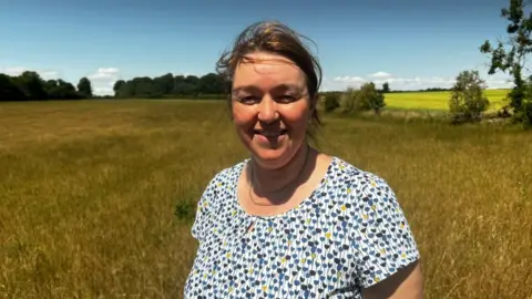 Nicola Cannon, a woman with tied-up hair and a patterned white and blue top, is stood in a field with trees on the horizon.