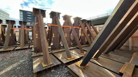 The rust coloured triangular steel props are lined up on ground near to the flyover, which can just be seen in the background. Each prop has yellow letters and numbers on the main, vertical central beam with two thinner props either side of the central section to create the triangle. They are each on top of pallets. In the background are two hi-rise buildings.