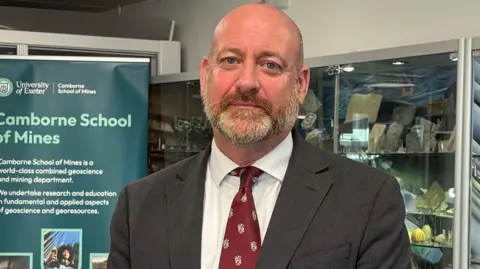 A man wearing a suit and tie looks towards the camera. He arms hanging down in front of him. He is standing indoors. A large sign behind him reads Camborne School of Mines.