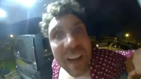 Cumbria Police Daniel Lytollis filmed by a doorbell camera. He is wearing a red shirt which is unbuttoned revealing his chest. He has brown messy hair and is staring wide-eyed into the camera.