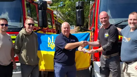 Five people stood in front of two fire engines and a Ukranian flag. Two of the people are shaking hands.
