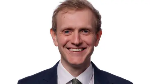 Joe Martin smiling at a camera wearing a dark suit and a white shirt, as well as a green tie