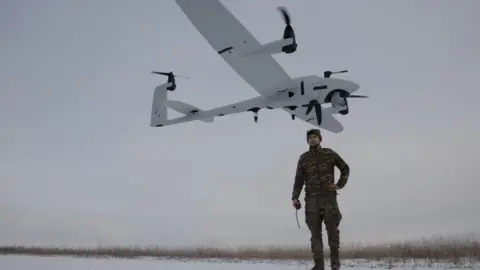 A Ukrainian serviceman in camouflage uniform stands on snow-covered ground after launching a reconnaissance unmanned aerial vehicle seen above his head