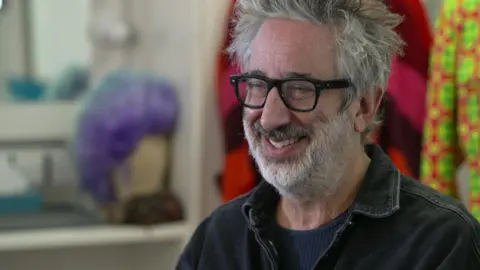 A man with black rimmed glasses and white hair laughs off camera