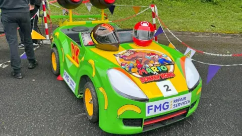 Hull 4 Heroes A bright green go-kart with yellow accents, designed to look like Donkey Kong's kart from Mario Kart. It has two helmets on the hood and large red rocket-like structures on the back. It's parked outdoors in a car park. People are stood nearby.