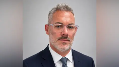 Deaf Connexions A head shot of Matthew Talbot. He has short grey hair and a beard and moustache. He is wearing a dark blue suit, white shirt and blue tie. 