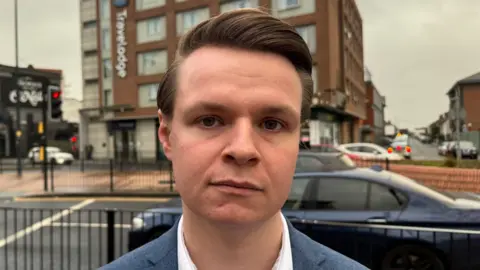 A portrait photo of Joshua Reynolds MP wearing a white shirt and a blue blazer with Maidenhead Travelodge seen in the background.