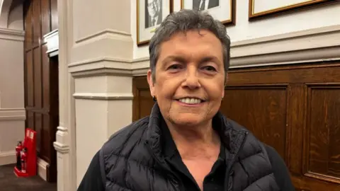 A woman wearing a black jacket stands in front of wooden paneling. She has short greying black hair and earrings.