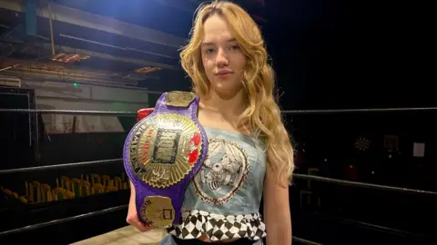 Zoe Dennis/BBC Annabelle with long blonde hair holding her purple and gold European Bellatix belt over her right shoulder. 