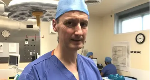 Mr Colbert stands in a hospital operating room, wearing blue scrubs and surgical hat. Two members of staff, also in scrubs, can be seen over his left shoulder looking at a screen.