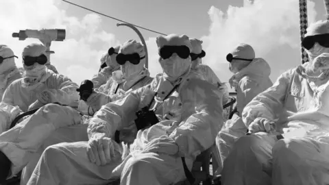 Getty Images Members of the press and the Royal Navy wearing white overalls and hoods and black goggles sit in seats on board a naval vessel ahead of nuclear tests in 1957