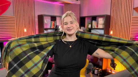 A blonde haired woman, Danni Diston, in a podcast studio with a large Cornish tartan scarf around her