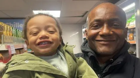 A bald man holds a smiling toddler in front of a supermarket fridge