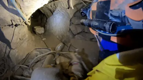 There is a tunnel and a plastic container on a string. Someone is pulling the string and you can see the back of their caving helmet. It is underground.
