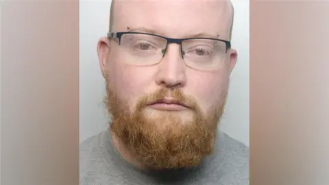 A mugshot of a man with glasses, a nose piercing, a ginger beard and a grey top.