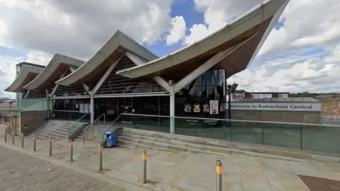 The front of Rotherham Central station, a modern building with sweeping, curved roof sections and large glass walls. Steps and a ramp lead up to the entrance, and a sign on the right reads “Welcome to Rotherham Central”. The area in front is paved with stone and lined with metal bollards.