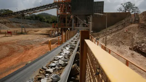 Reuters A conveyor belt carried a mixture of rocks and fine particles to a central building in the background. There is a yellow barrier to the right of the belt. Two workers in the background sort through the rocks. The surroundings are orange dusty ground with a few trees