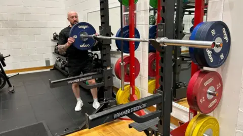 Lance Corporal Ceri Williams adds a 20kg weight to a barbell in the Tidworth Camp gym. He's wearing black t-shirt and shorts and white trainers. He has a brown beard.