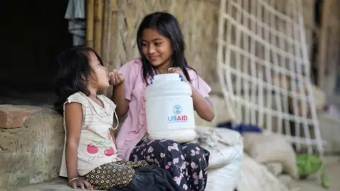 Reuters A Rohingya girl feeds a child from a jar with the USAID logo on it, at a refugee camp in Cox's Bazar, Bangladesh, on 11 February 2025.