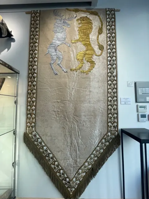 This image shows a large tapestry featuring the sigils of House Baratheon and House Lannister. A silver stag and a golden lion are depicted standing up, bordered with gold embroidery and fringe at the bottom.