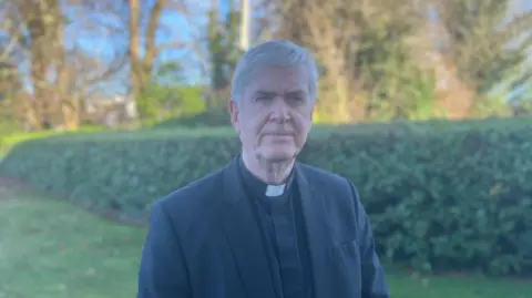 A priest is looking at the camera. He's standing on grass. He has grey hair. He has a neutral expression. He's wearing his black priest suit with a white collar. There are bushes and trees behind him. 