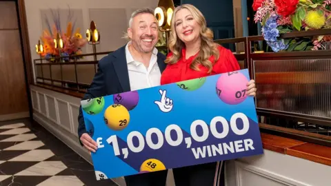 Richard Davies and Faye Stevenson-Davies hold a sign showing lottery balls and text saying one million pounds winner.
