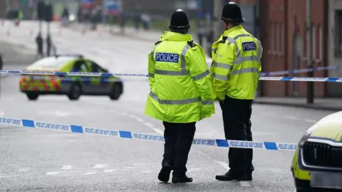PA Media Two officers stand with their backs to the camera next to police tape with a police car in the road in the background in Leicester city centre