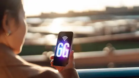 Someone looking at their smart phone which says 6G in big letters. They can sat down and their background is blurry.