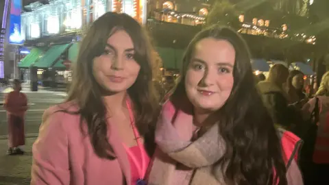 BBC two women stand stand outside City Hall. The women on the left has brown hair, wears a pink blazer with a pink top underneath. The other has on a Pink and grey scarf with a pink hi-viz jacket, she has brown hair